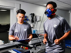 A student using a treadmill while wearing a breathing apparatus, being monitored by another student