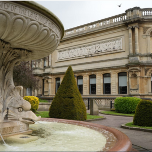 Museum in Wolverhampton captured from the nearby park, numerous windows and sculpted patterns visible past the park bushes and fountain