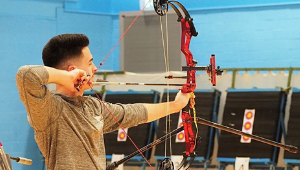 A student practicing archery, drawing back a large bow in front of a target range