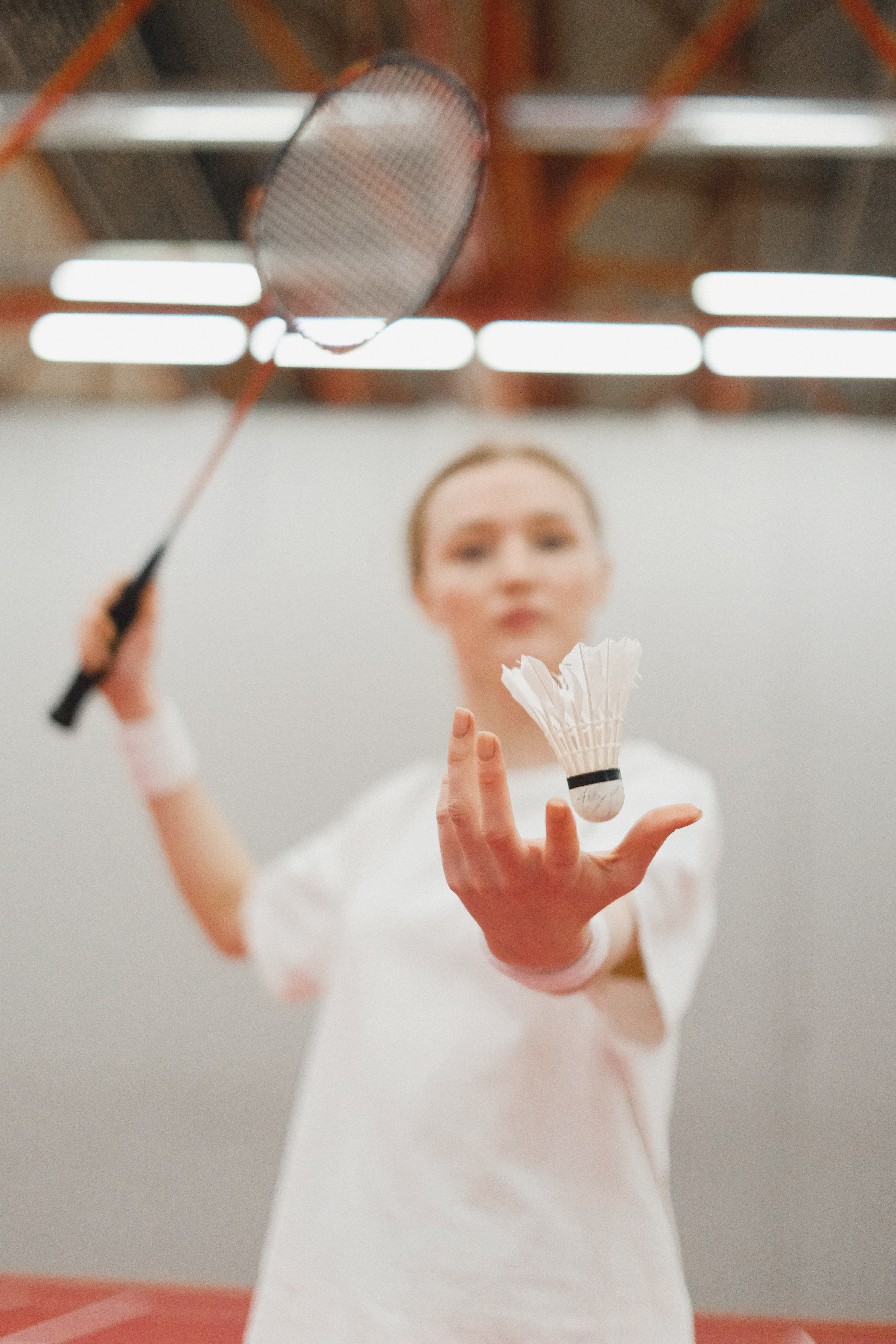 A badminton player throwing the shuttlecock and pulling back her racket in preparation to serve, playing on an indoor court