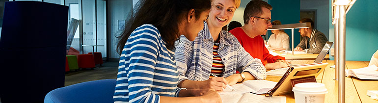 Students studying at a desk, conversing and working on paper and tablets