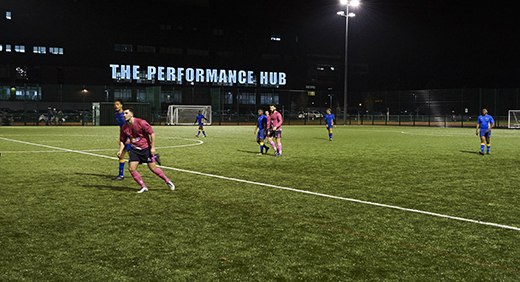 Football played outdoors at night on a Walsall Campus outdoor pitch, the performance hub signage lit up in the background